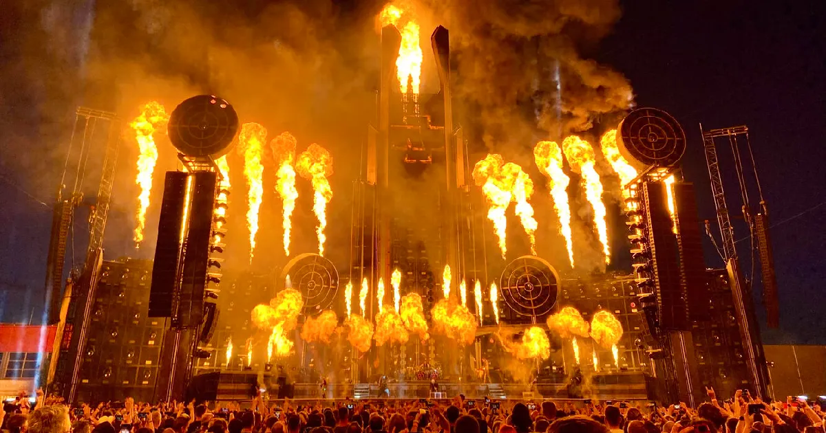 Rammstein performing on a massive industrial-style stage as towering flames and bursts of pyrotechnics erupt behind them, casting dramatic orange light over the band and the crowd.