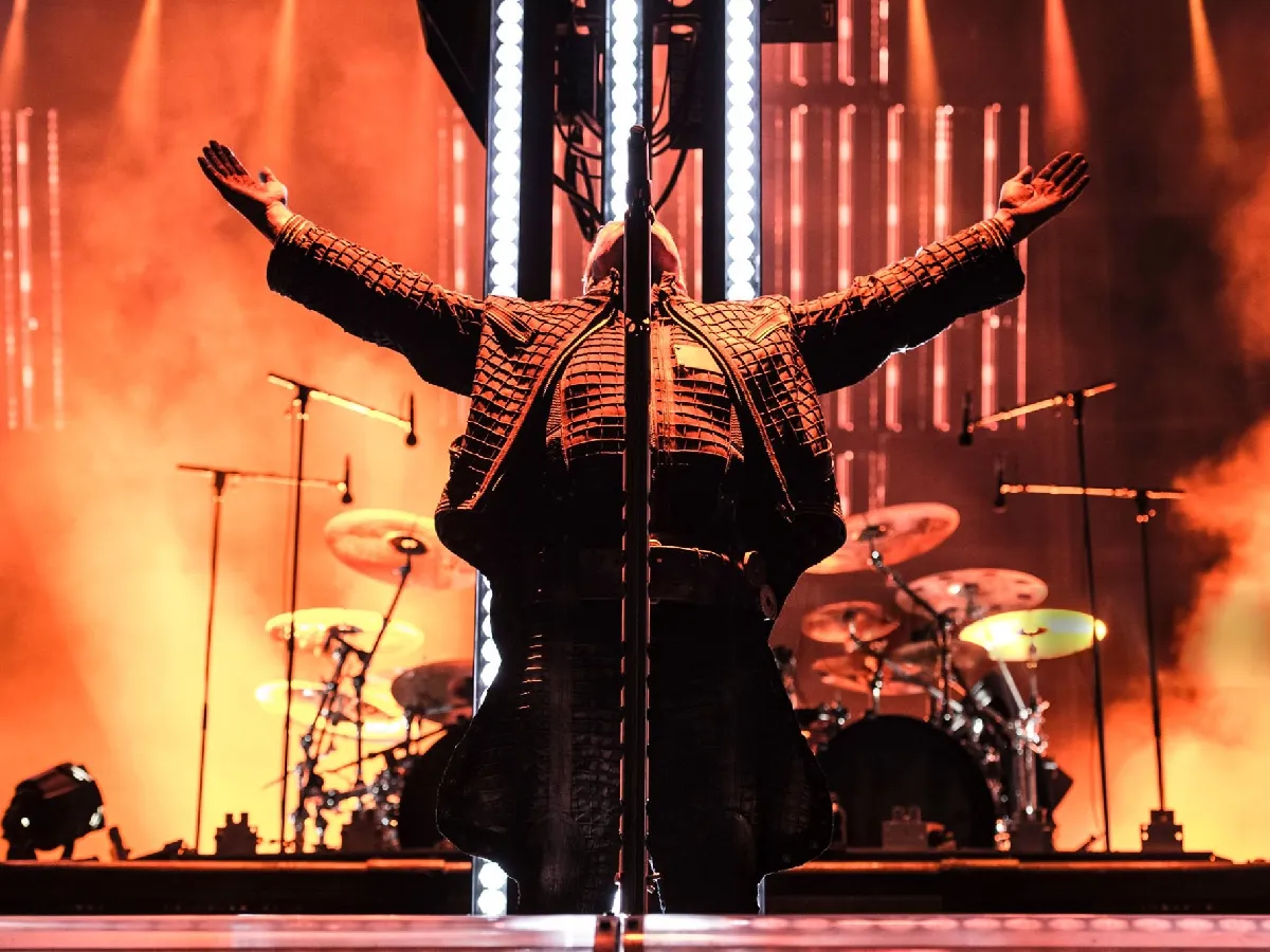 Till Lindemann performing live on stage with the rest of Rammstein, surrounded by industrial lighting and dramatic stage effects.